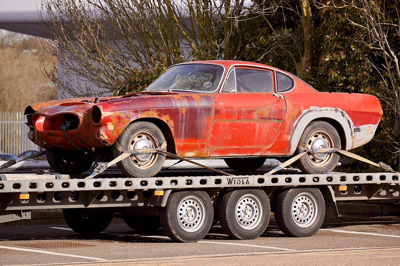 A worn-out, rusty vintage car secured on a flatbed trailer, outdoors under daylight.